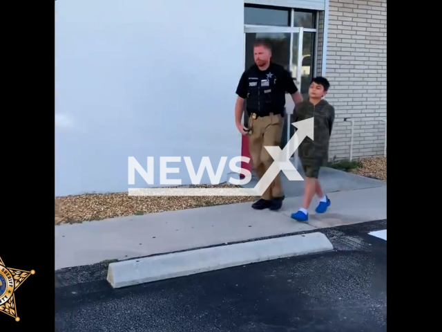 Policeman escorting the 5th grader that was arrested for sending a text message threatening to conduct a mass shooting, on the 29th of May, in Cape Coral, Florida. Note: This picture is a screenshot from the video (Lee County Sheriff's Office/Clipzilla).