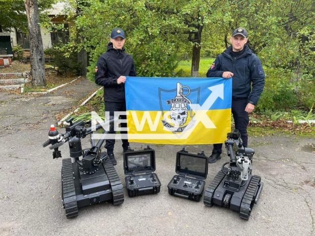 Two Ukrainian sappers hold Ukrainian flag near demining robots in Vinnytsia, Ukraine in undated photo. Sappers of the State Emergency Service of Ukraine received two modern American robots for remote demining. Note: Photo is obtained from the State Emergency Service of Ukraine (@MNS.GOV.UA/Newsflash)